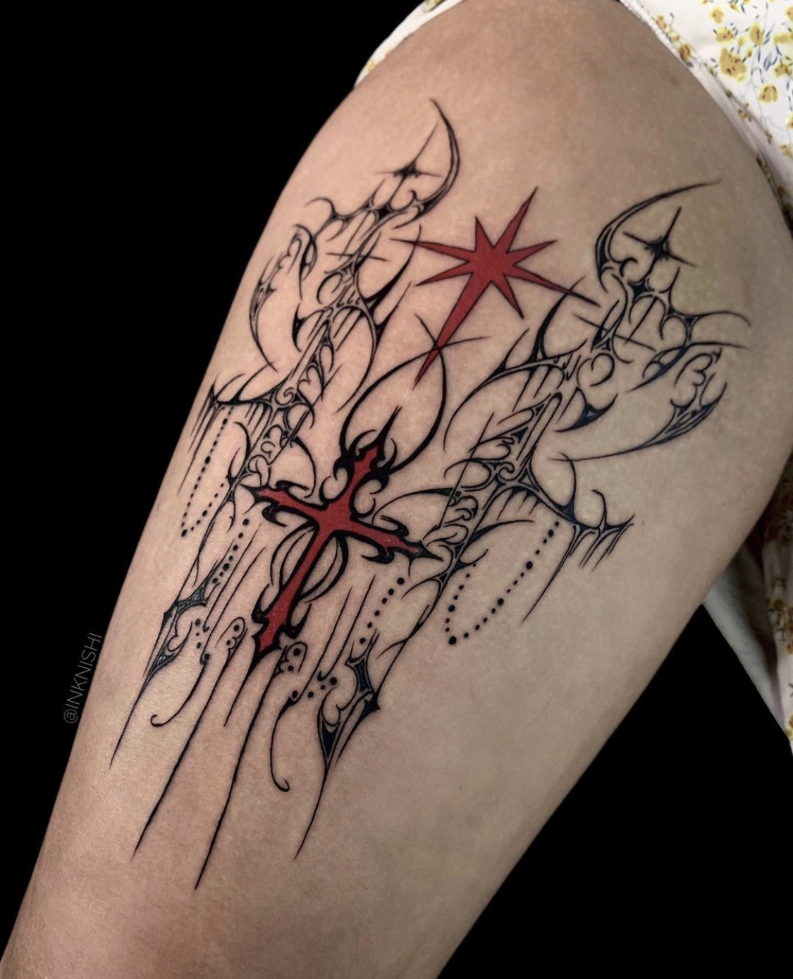 Tatuagem ornamental detalhada na coxa, com linhas pretas finas e elementos pontiagudos, representando uma composição mística. No centro, uma cruz estilizada em vermelho, rodeada por formas curvas que se estendem elegantemente, criando um contraste marcante com os detalhes pretos. Acima da cruz, uma estrela vermelha de oito pontas adiciona um toque celestial ao design, que combina elementos góticos e simbólicos.